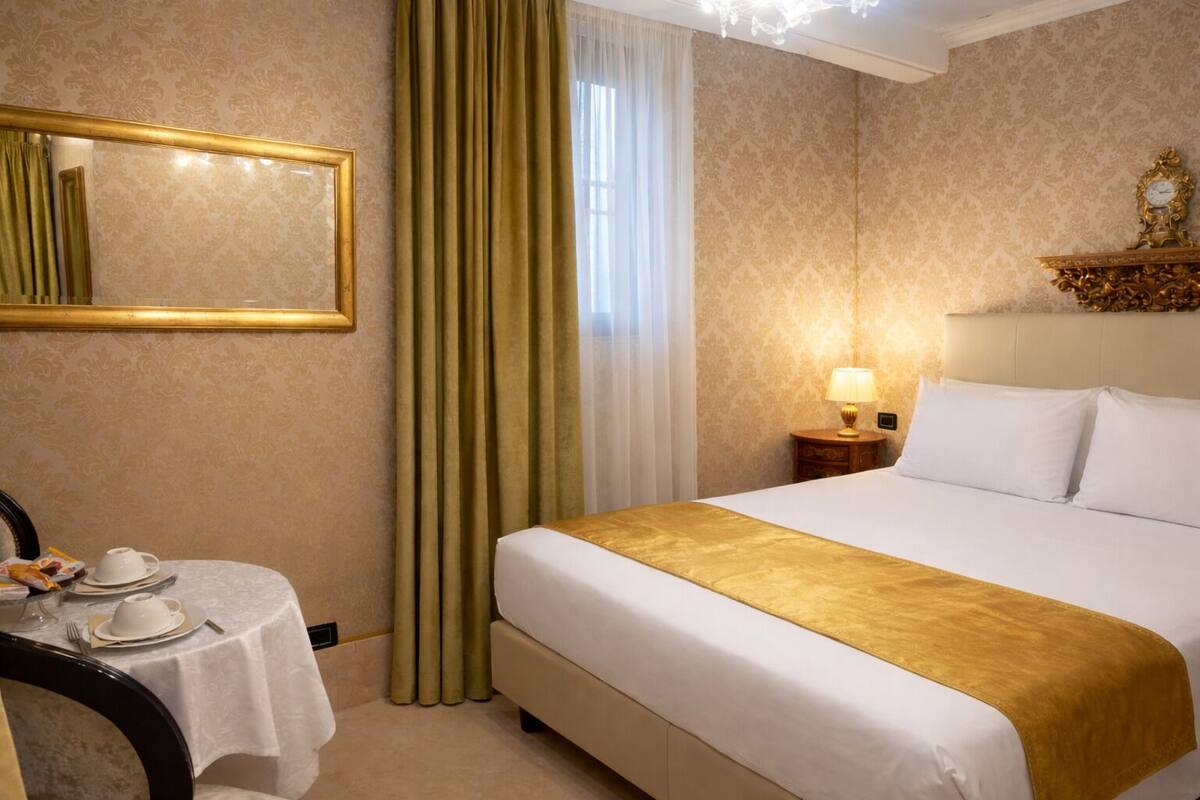 Una habitación de hotel con una cama, una mesita de noche con lámpara, un espejo y una pequeña mesa con una taza y un plato.