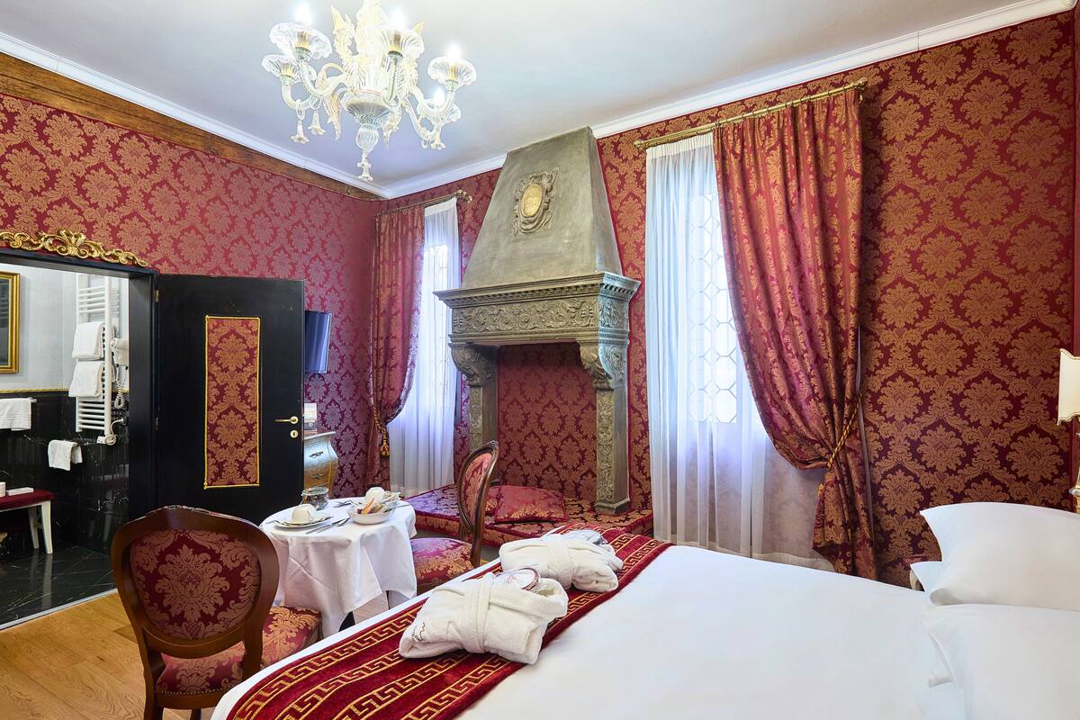 Una habitación de hotel con una cama grande, una silla de madera, una mesa con mantel individual rojo y dorado, y una chimenea.