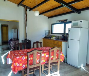 A compact kitchen with a refrigerator, microwave, and a small window. A dining table with chairs is positioned under a wooden beam ceiling. - Arriba del Valle - Casas de Altura (Potrerillos)