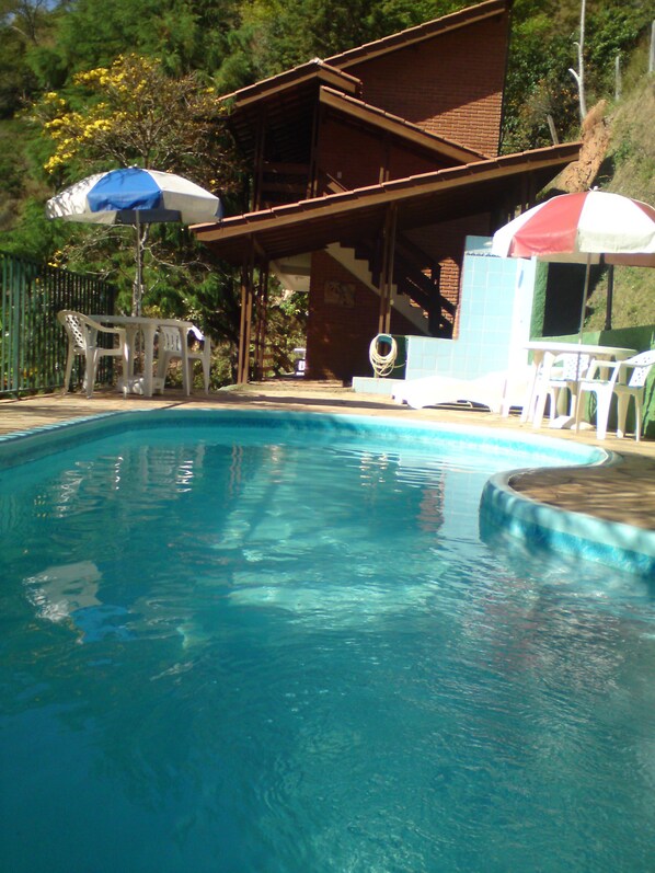 A swimming pool with a wooden deck, surrounded by white plastic chairs and umbrellas. - Chalés Pousada Serra Negra (Serra Negra)