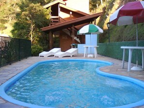 A swimming pool with lounge chairs and a table, surrounded by a fenced area and a building with a staircase. - Chalés Pousada Serra Negra (Serra Negra)