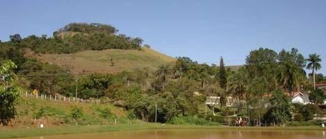 A body of water with a hill in the background, surrounded by greenery and a few buildings.
