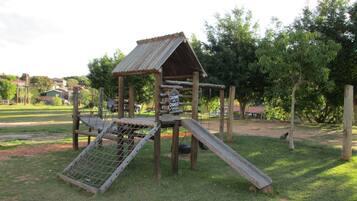 Une structure de jeux en bois, dotée d’un toboggan et d’un filet d’escalade, entourée d’herbe et d’arbres.