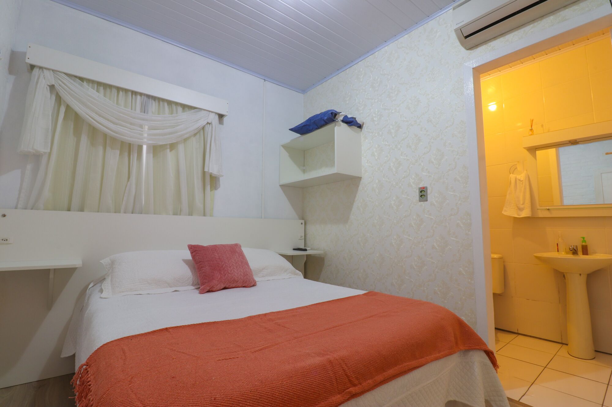 Habitación de hotel con cama, mesita de noche, espejo, baño con lavabo e inodoro, y ventana con cortinas.