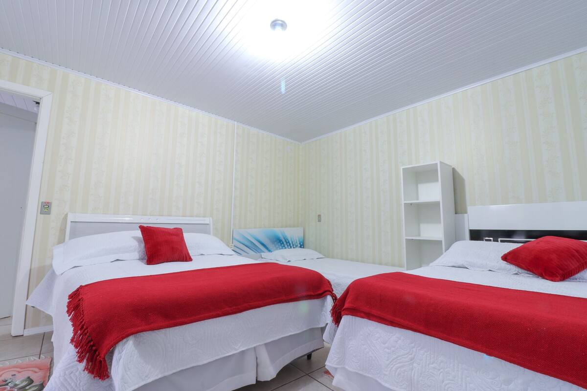Une chambre avec deux lits, chacun recouvert d’une couverture rouge et doté de draps blancs. On y trouve une tête de lit blanche, un tableau au mur et une étagère blanche.