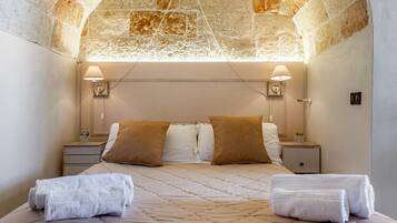 A bedroom with a stone ceiling, two bedside tables with lamps, a bed with a quilted cover, and two towels on the bed.