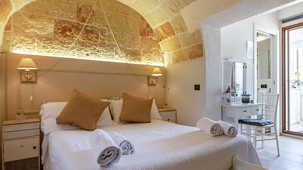 A hotel room with a stone arch ceiling, a large bed with white bedding and brown pillows, a bedside table with a lamp, and a chair in the corner. - Respiro del Mare (Polignano a Mare)