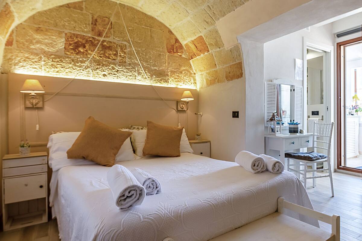 Habitación de hotel con techo abovedado de piedra, una cama grande con ropa de cama blanca y almohadas marrones, una mesita de noche con una lámpara y una silla en la esquina.