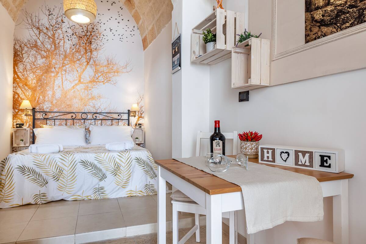 Un dormitorio con cama, mesitas de noche, una mesita auxiliar, una mesa de madera con un cartel que dice 'HOME' y una estantería de pared con adornos.