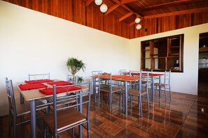 Un comedor con sillas y mesas de metal, piso de madera, un ventanal grande y un techo con vigas de madera y luces colgantes.