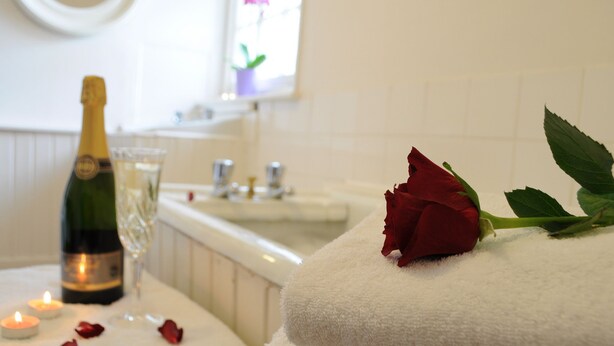 Une salle de bain avec une baignoire blanche, une bouteille de champagne, un verre et une rose rouge posée sur une serviette.