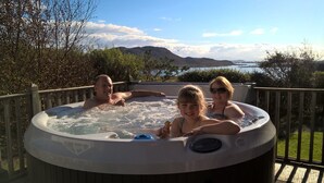 A hot tub with three people, surrounded by a wooden deck and a scenic view of mountains and a lake. - Windyhill Lodge 14665 (Ullapool)