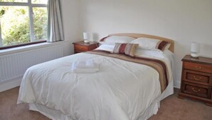 A neatly made bed with a white duvet and a brown and white striped blanket, a wooden nightstand with a lamp, and a window with curtains. - Surima, Chichester 48663 (Chichester)
