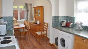 A compact kitchen with wooden cabinets, a built-in washing machine, a dining table with chairs, and a window with curtains. - Surima, Chichester 48663 (Chichester)