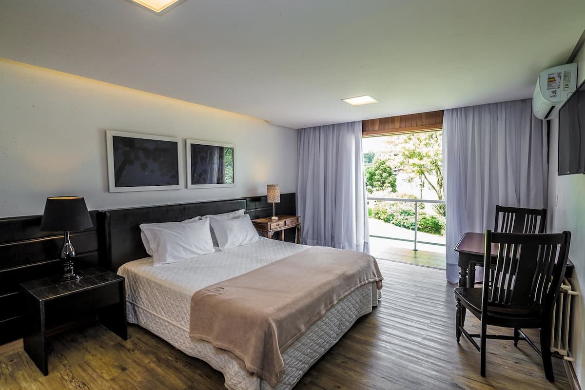 A hotel room with a bed, a nightstand, a chair, a table, and a window with curtains.