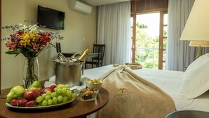 Habitación de hotel con cama, una mesa de comedor con frutas y una botella de champán, televisor y ventana con cortinas.