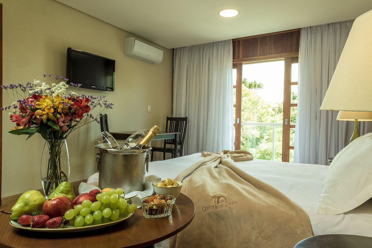 A hotel room with a bed, a dining table with fruits and a bottle of champagne, a TV, and a window with curtains.