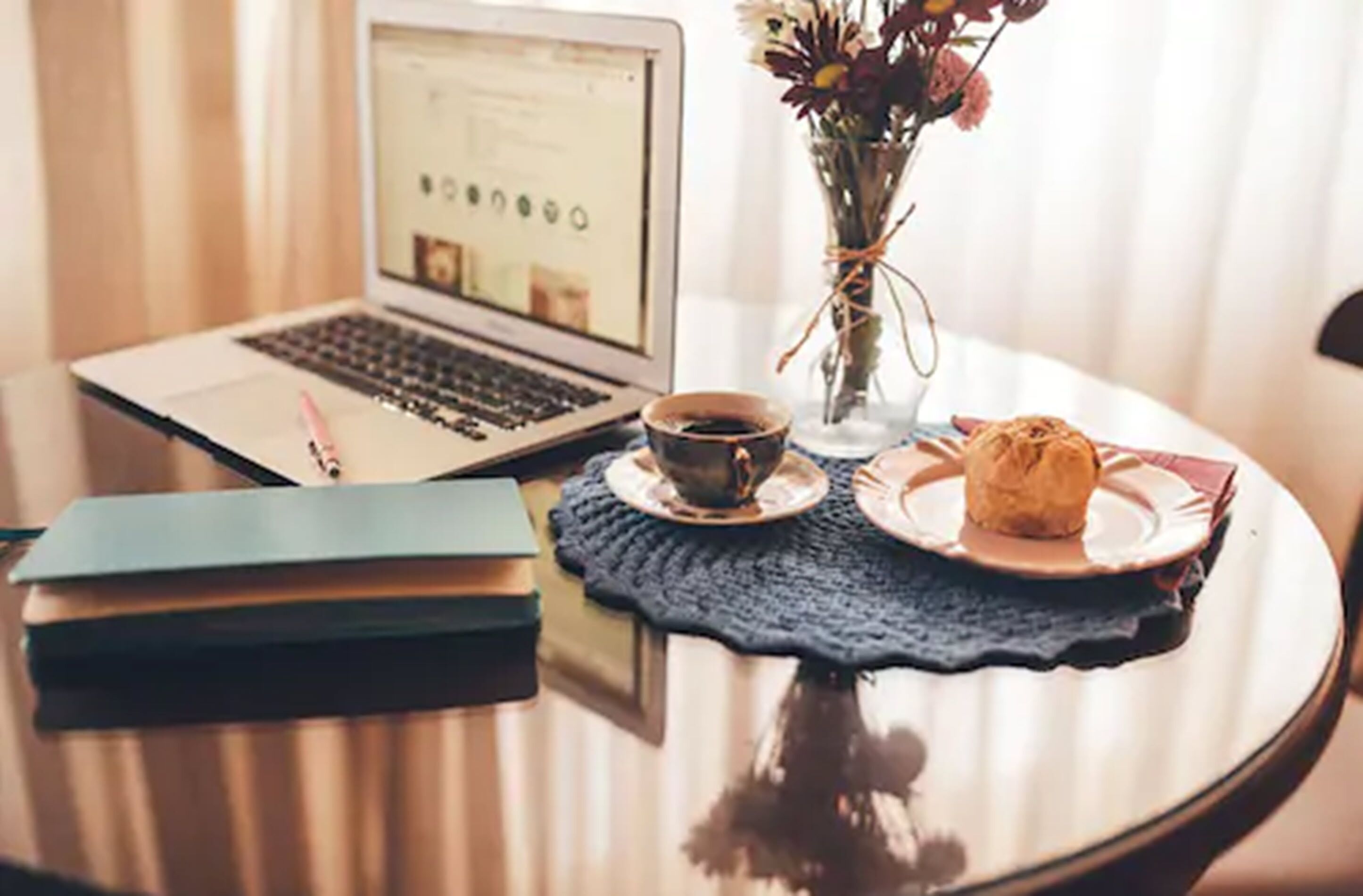 A laptop on a table with a cup of coffee, a plate with a pastry, and a vase with flowers.