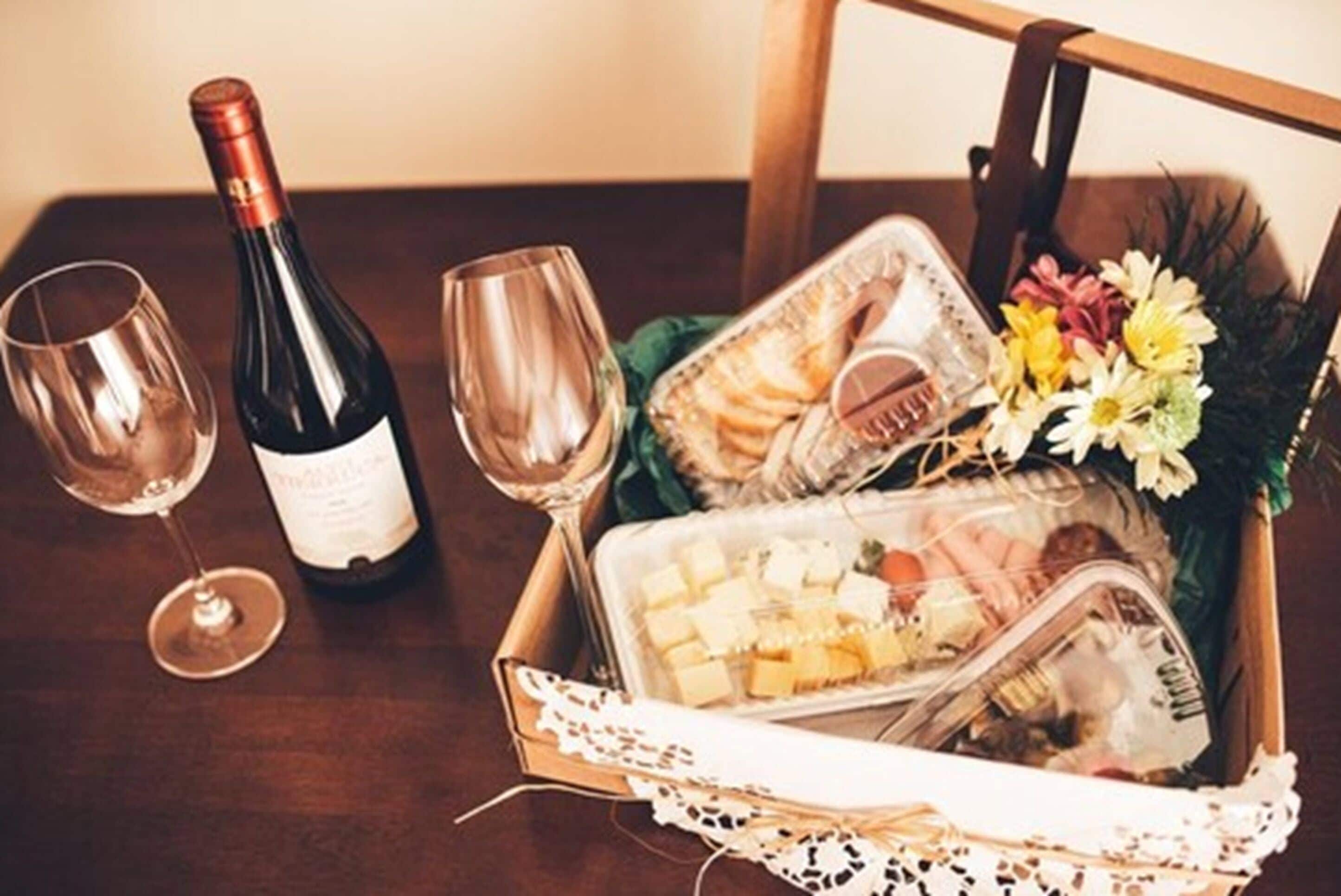 Una mesa de madera con una botella de vino, dos copas de vino, una cesta con queso, galletas, flores y una revista.