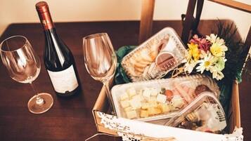 Una mesa de madera con una botella de vino, dos copas de vino, una cesta con queso, galletas, flores y una revista.