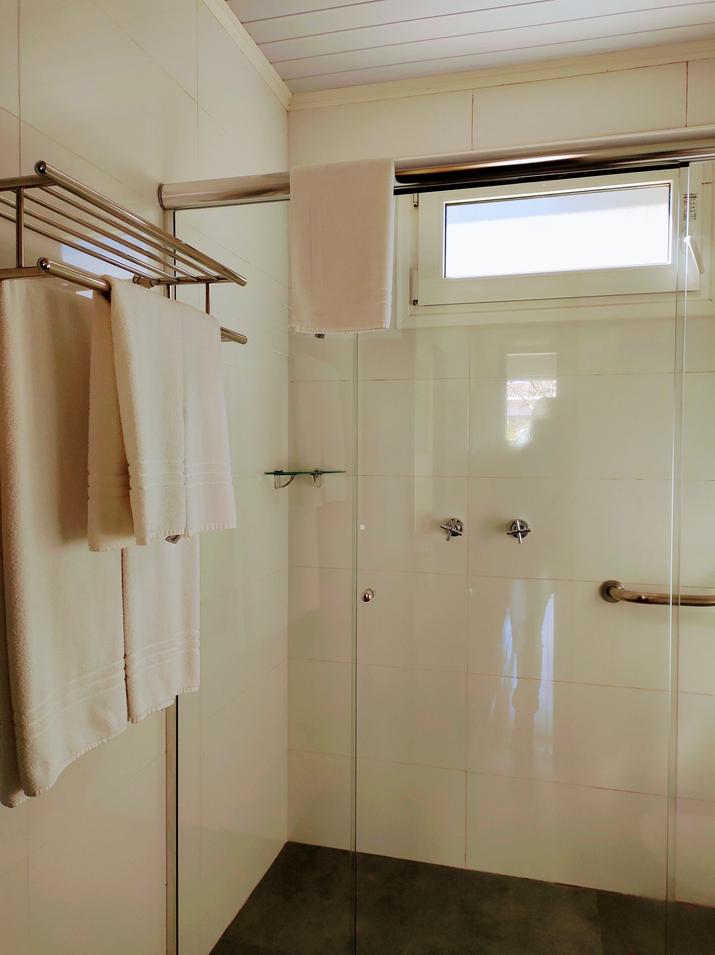 Un baño con cabina de ducha de vidrio, un perchero con varias toallas y una ventana sobre la ducha.