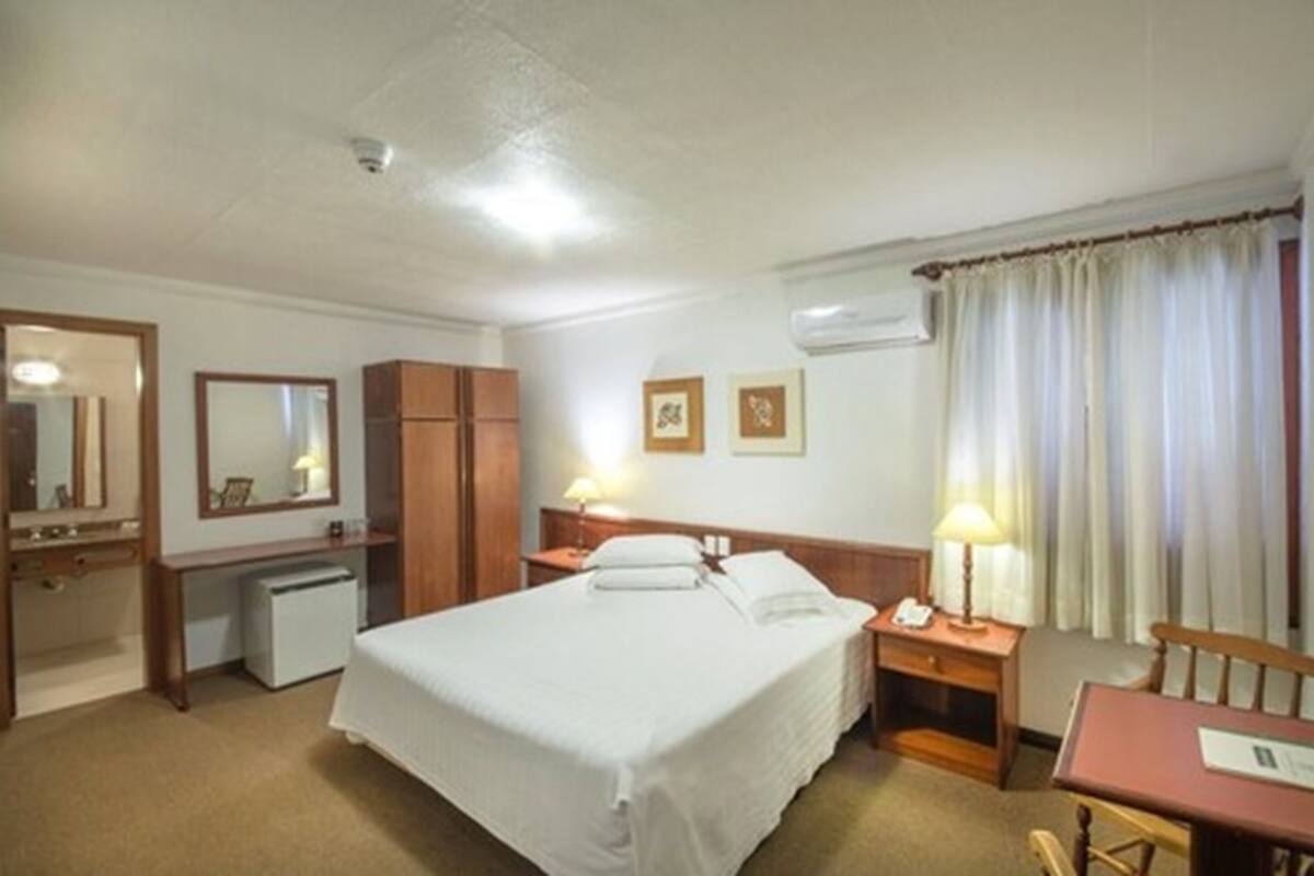 A hotel room with a bed, bedside table, lamp, desk, chair, and a small refrigerator.