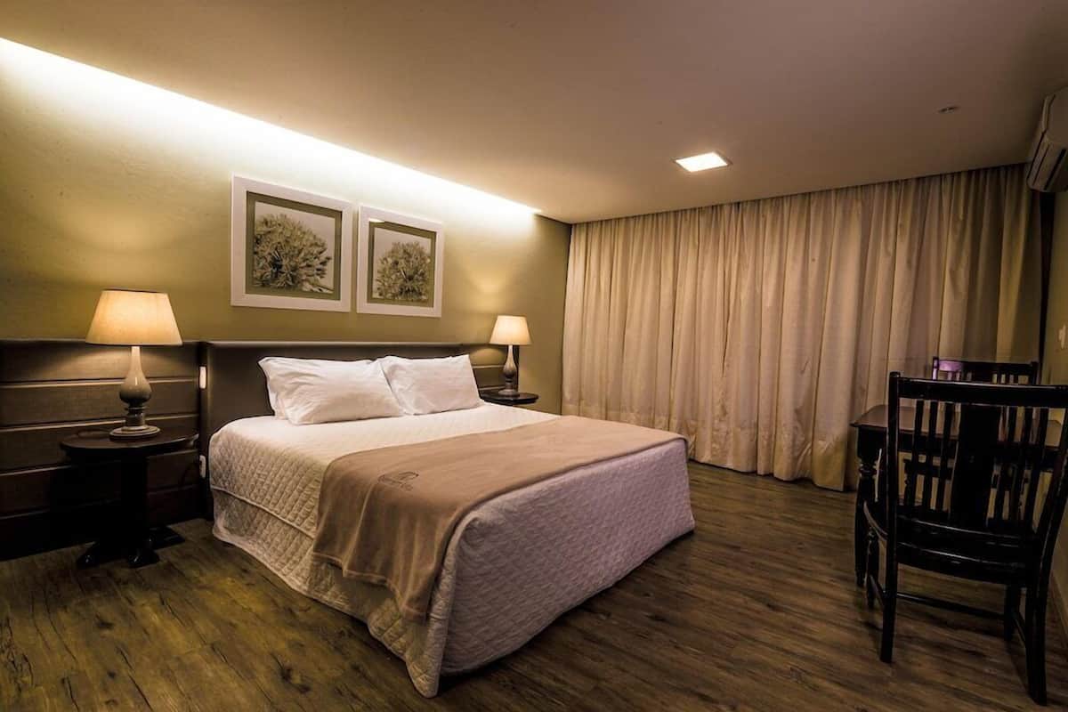 A hotel room with a bed, two bedside lamps, a chair, a desk, and a large window with curtains.