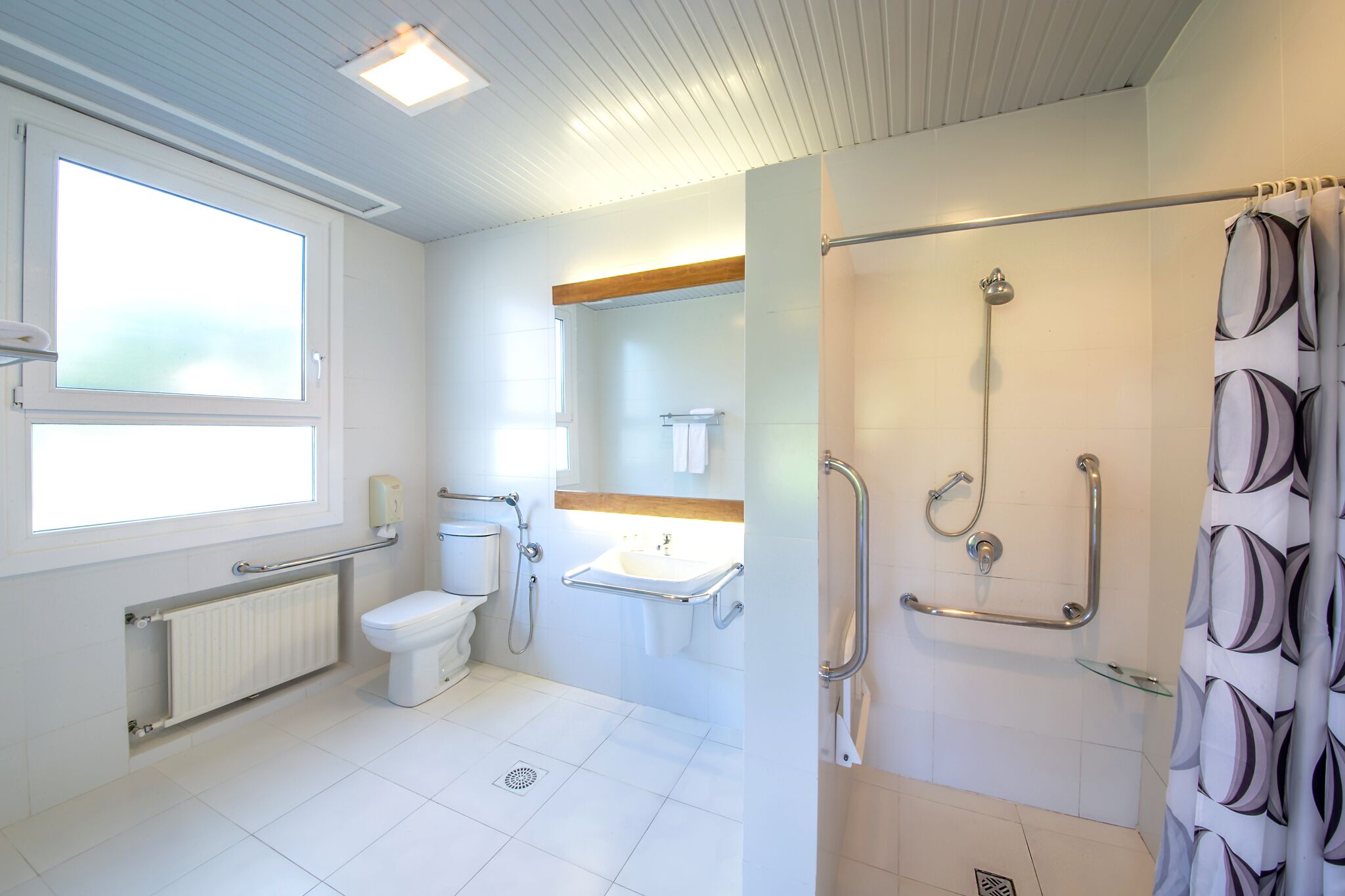 A modern bathroom with a toilet, sink, and shower. It includes a window, a towel rack, and a handrail for accessibility.