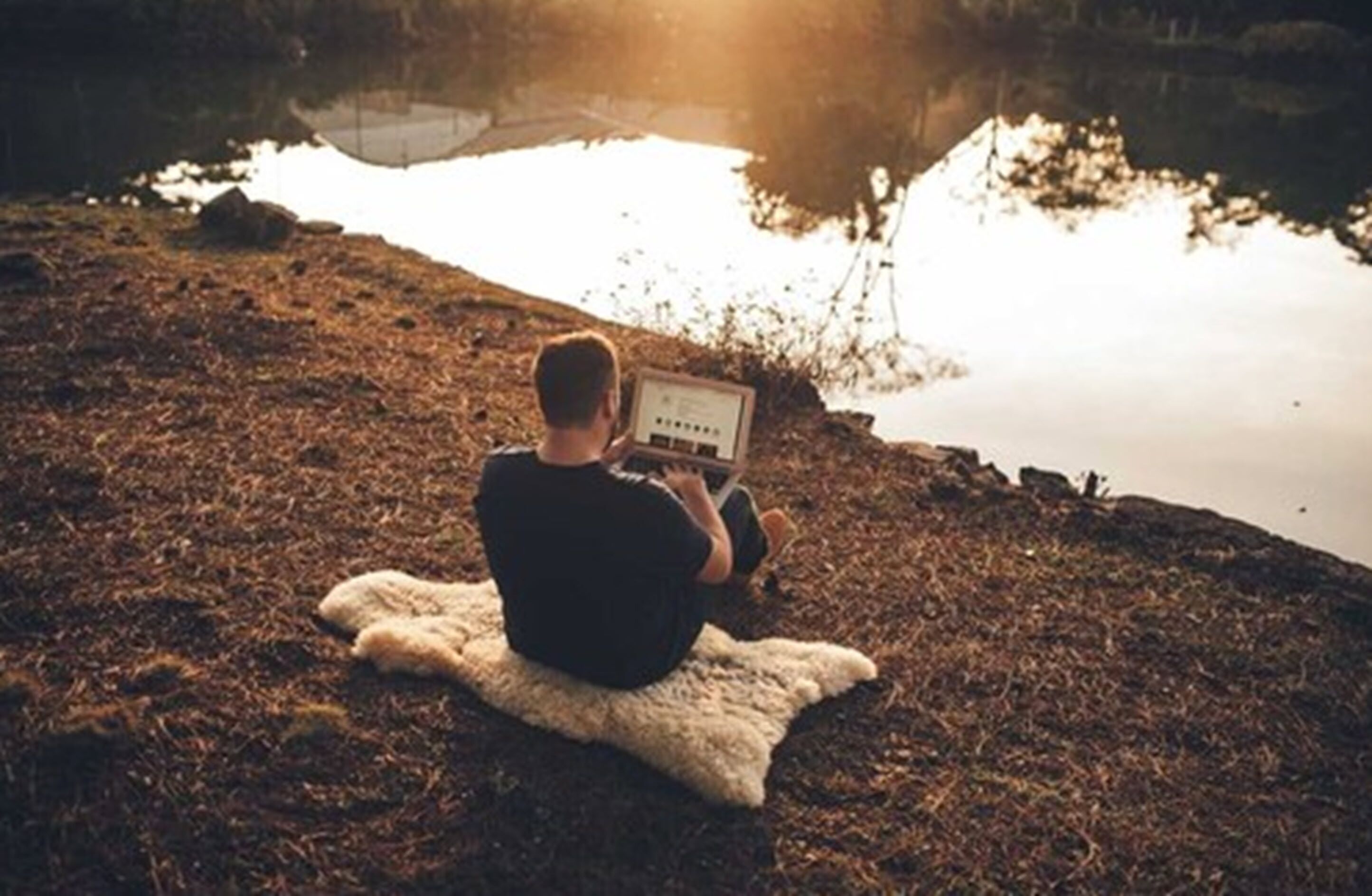 A person sitting on a blanket by a body of water, using a laptop.