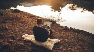 A person sitting on a blanket by a body of water, using a laptop.