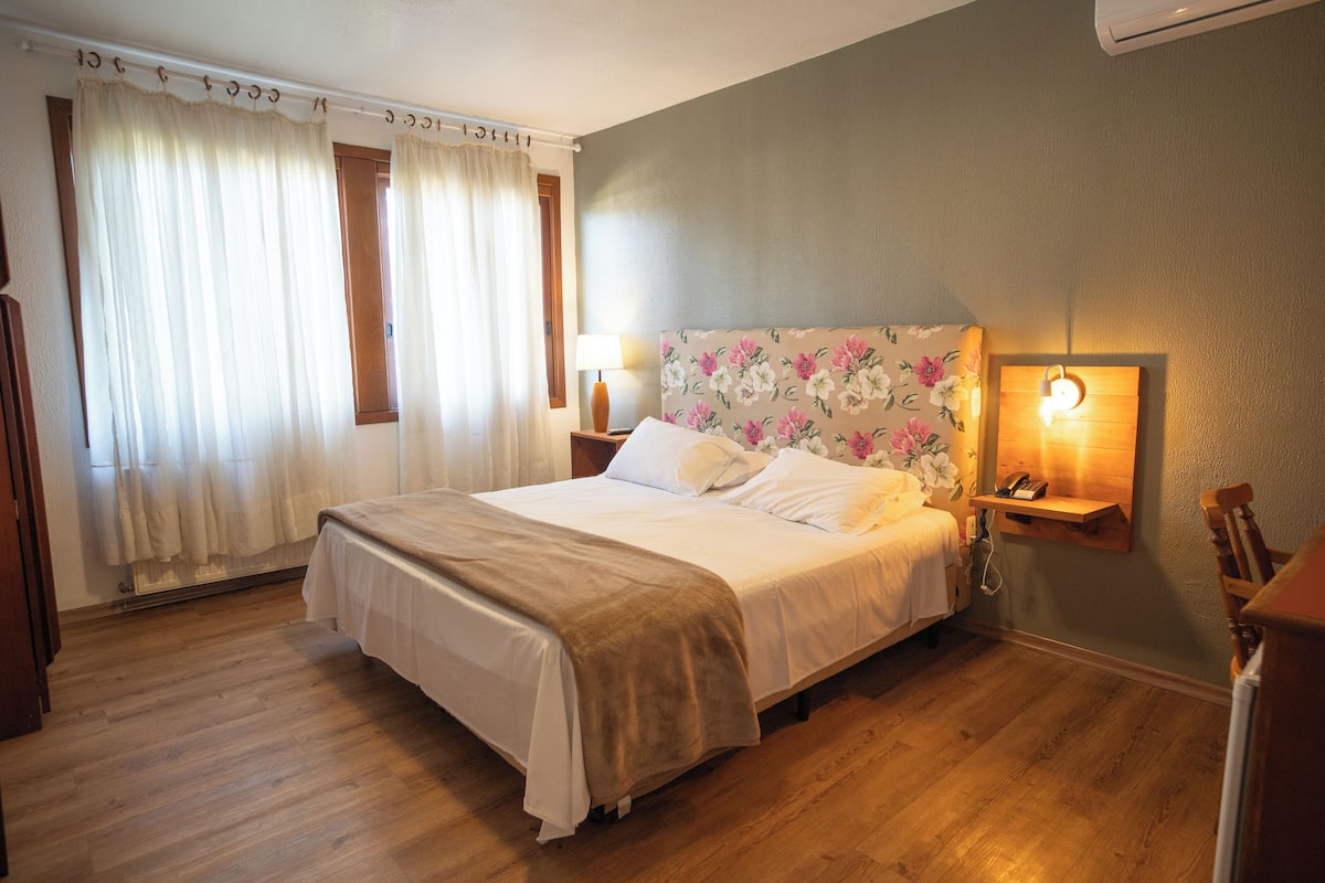 A hotel room with a bed, curtains, a nightstand with a lamp, a wall-mounted shelf with a phone, and a wooden chair.