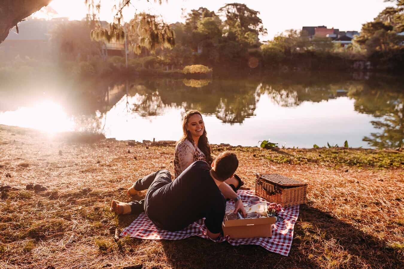 Una pareja descansando sobre una manta a cuadros junto a un lago, con una cesta de picnic y una botella de agua.