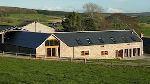 A stone building with a sloped roof and multiple windows, surrounded by a fenced property with a grassy foreground and rolling hills in the background.