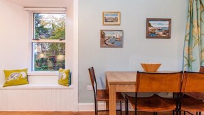 Uma área de refeições com mesa e cadeiras de madeira, janela com vista para árvores e duas pinturas emolduradas na parede.