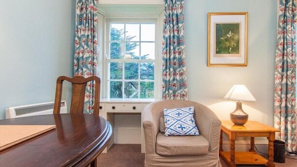 A room with a wooden chair, a cushioned armchair with a patterned pillow, a small table with a lamp, and a window with curtains. - Nursery (Isle of Colonsay)