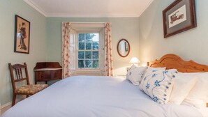 A bedroom with a large bed, a wooden headboard, a nightstand, a chair, a window with floral curtains, and two framed pictures on the wall. - Nursery (Isle of Colonsay)