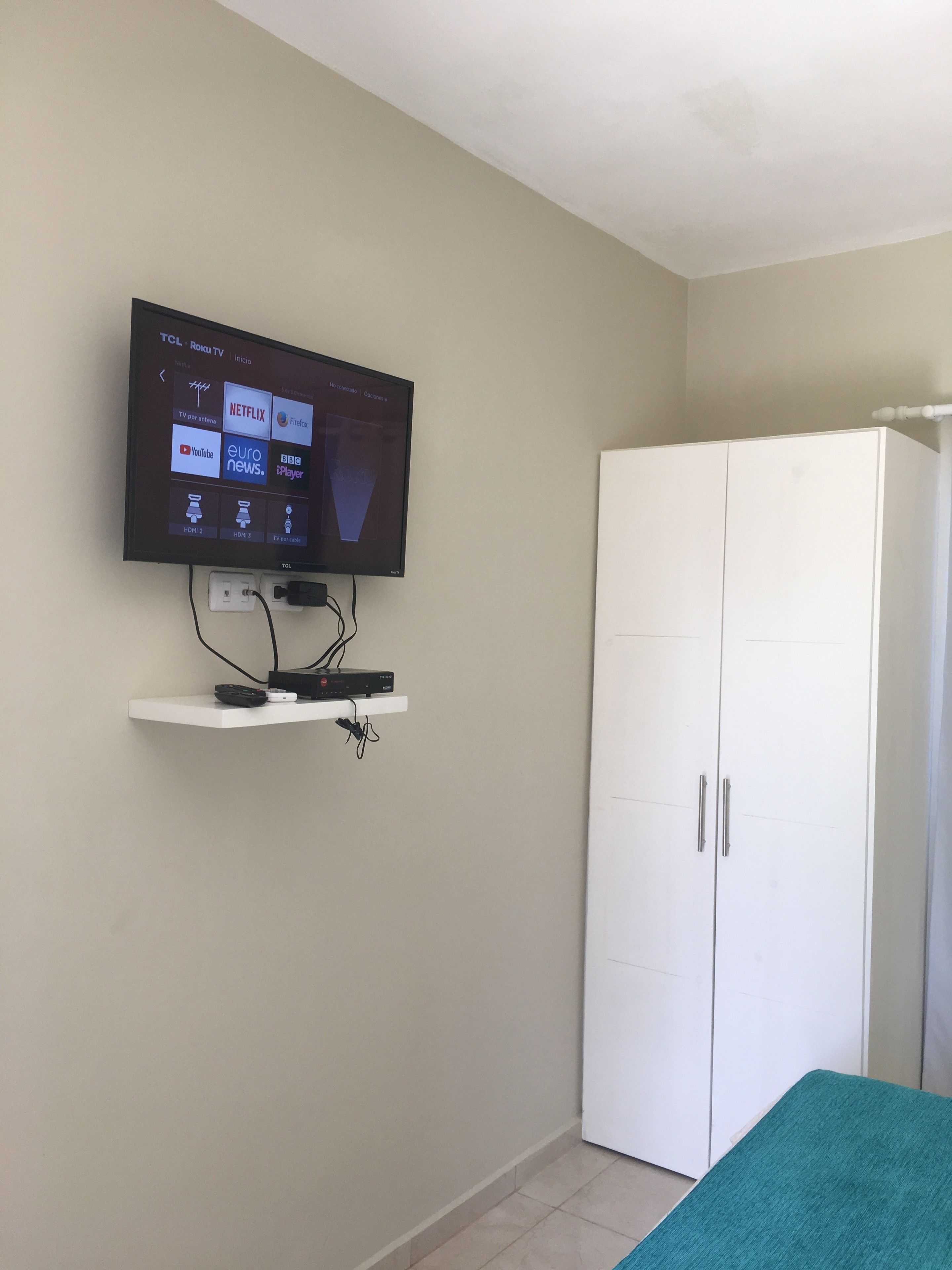Quarto com televisão montada exibindo diversos serviços de streaming, guarda-roupa branco e colcha de cama verde-água.