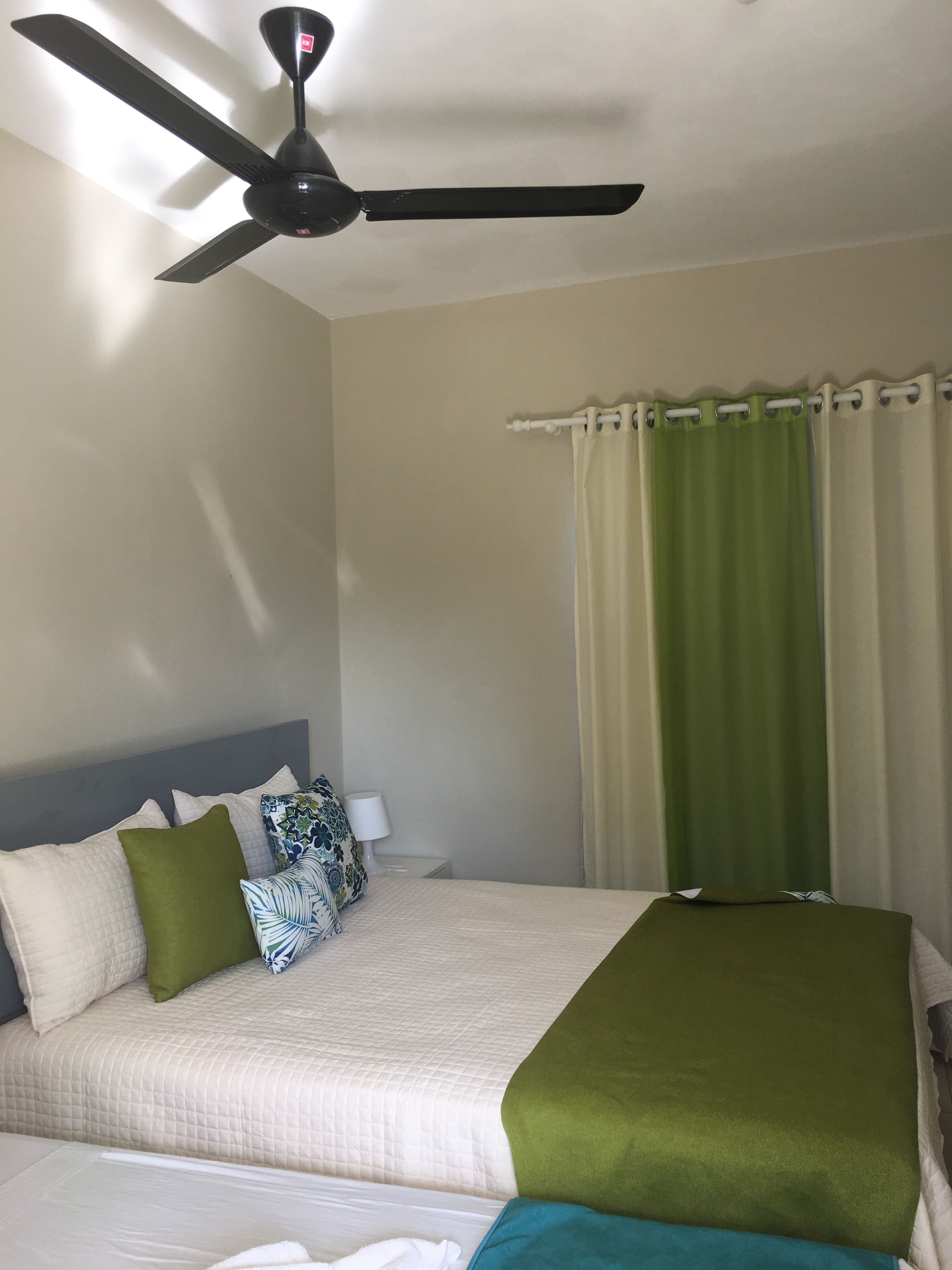 Camera d'albergo con un letto, una ventola a soffitto, un comodino con una lampada e una finestra con tende.