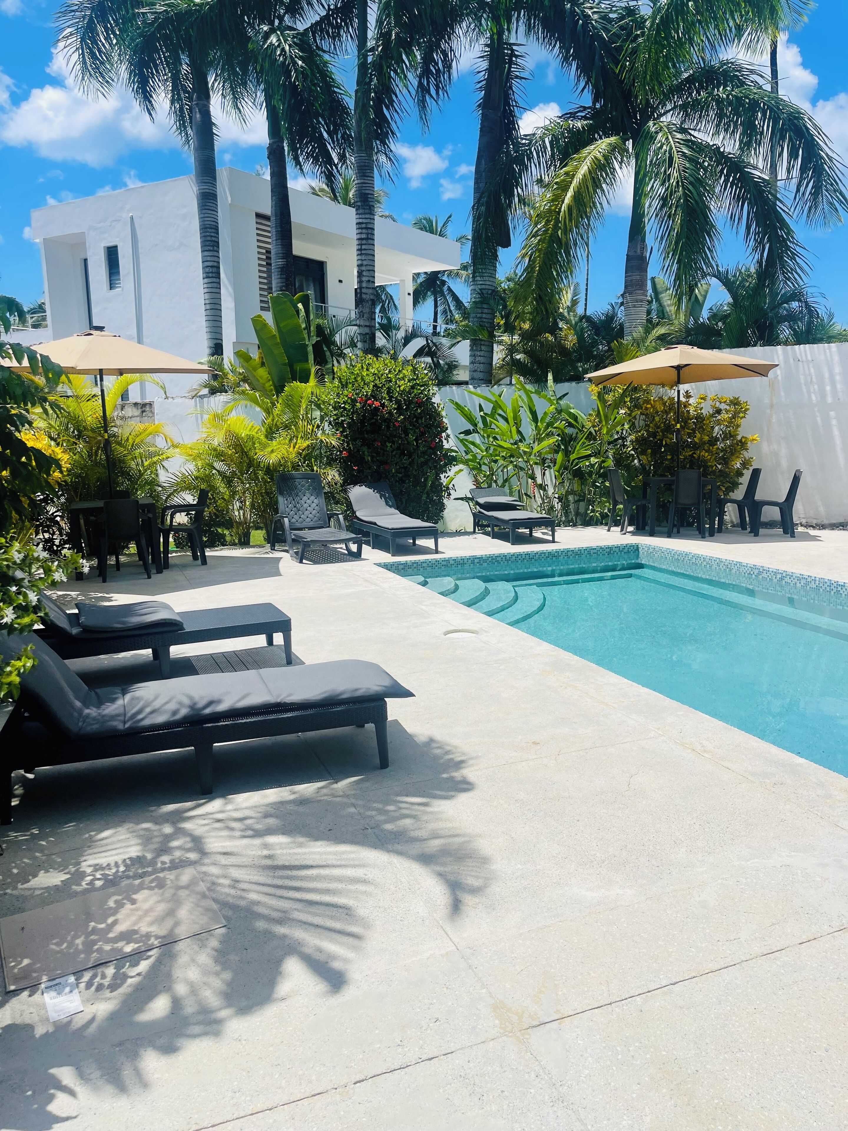 A pool area with lounge chairs and a table under a canopy, surrounded by palm trees and a white building.