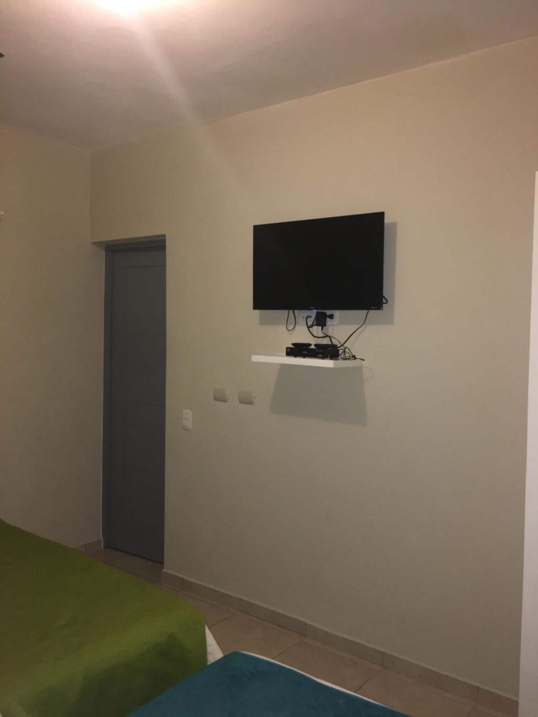 Una habitación con televisión montada en la pared, una puerta y una cama con colcha verde y azul.