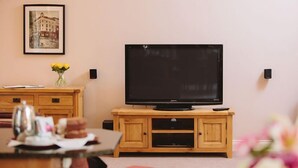 A living room with a flat-screen TV on a wooden stand, a framed picture on the wall, and a vase with yellow flowers on a wooden table. - Watson Cottage (Hot Tub) (Brampton)