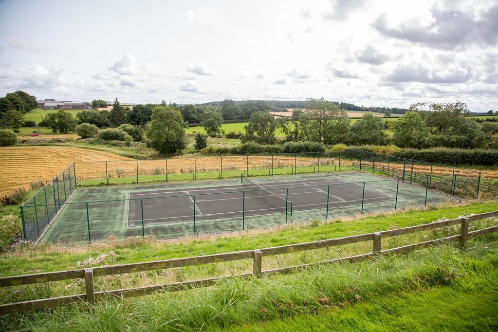A tennis court surrounded by a wooden fence, with a grassy field and trees in the background.