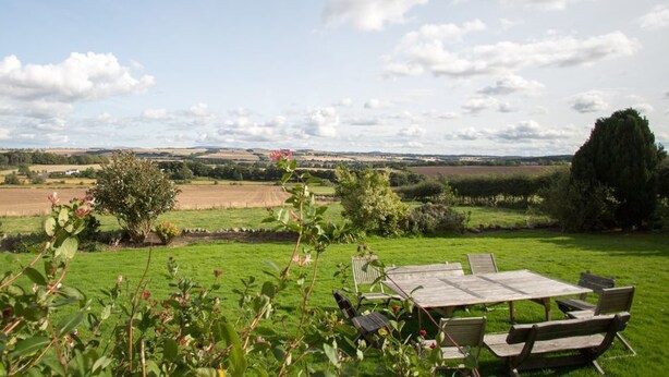 A garden with a wooden table and chairs, surrounded by greenery and a view of the countryside. - Edmondston House 163451 (Kelso)