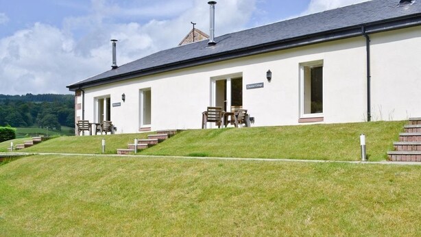 En byggnad med en vit fasad, en skorsten och en skylt med texten ”Chapel Cottage”. Det finns trappsteg som leder upp till entrén, och en bänk syns utanför.