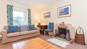 A cozy living room with a beige sofa, a blue armchair, a wooden coffee table, a fireplace, and a window with curtains. - Scalasaig Farmhouse (Isle of Colonsay)