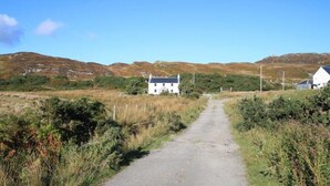 A rural road leading to a white house with a dark roof, surrounded by grassy fields and hills. - Scalasaig Farmhouse (Isle of Colonsay)