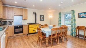 A compact kitchen with wooden cabinets, a microwave, and a stove. There is a dining area with a table and chairs, and a window with curtains. - Scalasaig Farmhouse (Isle of Colonsay)