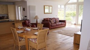 A dining area with a wooden table and wicker chairs, a leather sofa, and a large window providing a view of greenery.
