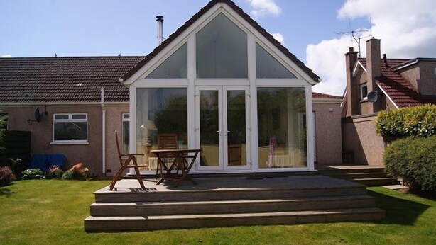 A house with a large glass conservatory, a patio with a table and chairs, and a well-maintained lawn.