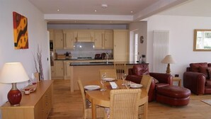 A modern living room with a dining table set for four, a kitchen in the background, and leather sofas.